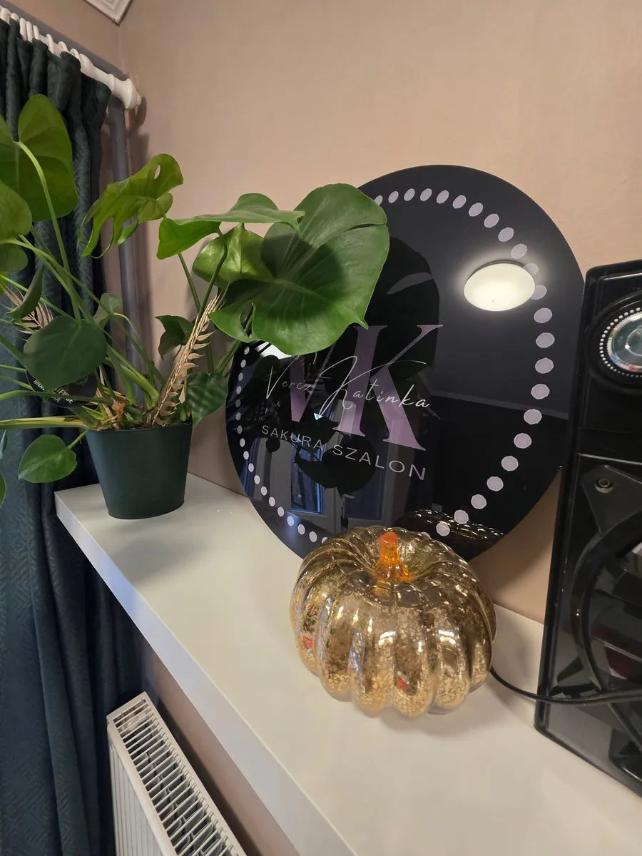 Sarok a Sakura Szalonban: kerek, fekete cégtábla a szalon monogramjával, egy monstera növény és egy arany üvegtök a fehér polcon.